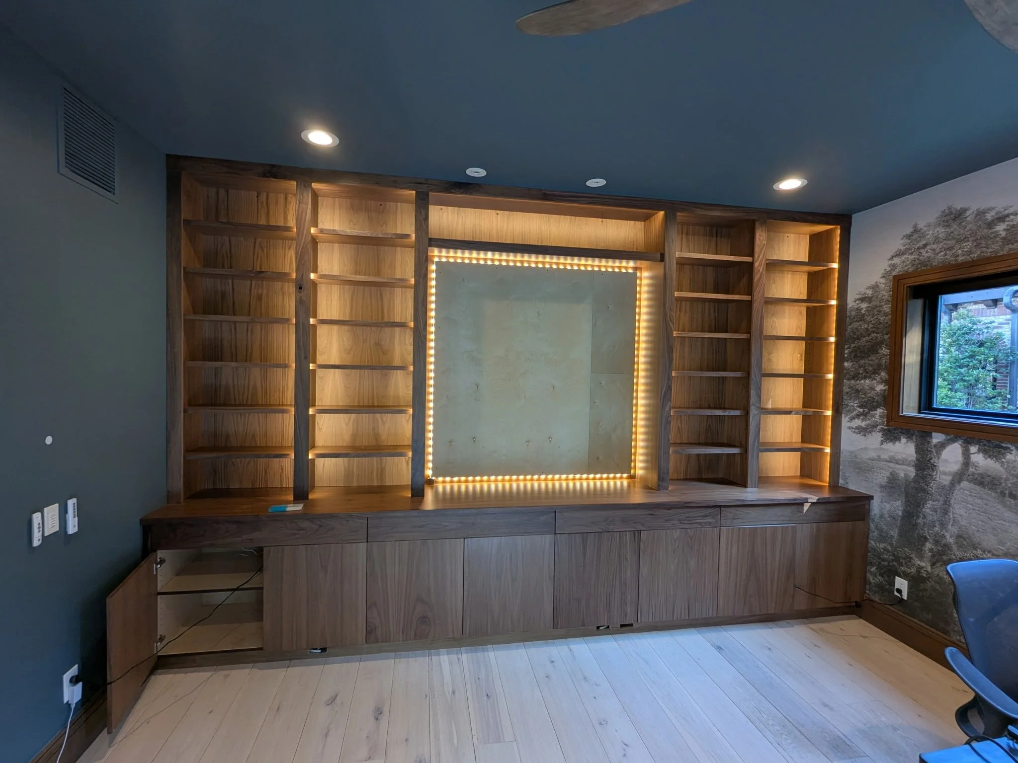 Empty wooden bookshelf with a built-in lighting feature in the center, located in a room with wood flooring, a window showing outdoor greenery, and a mural of trees on the wall.
