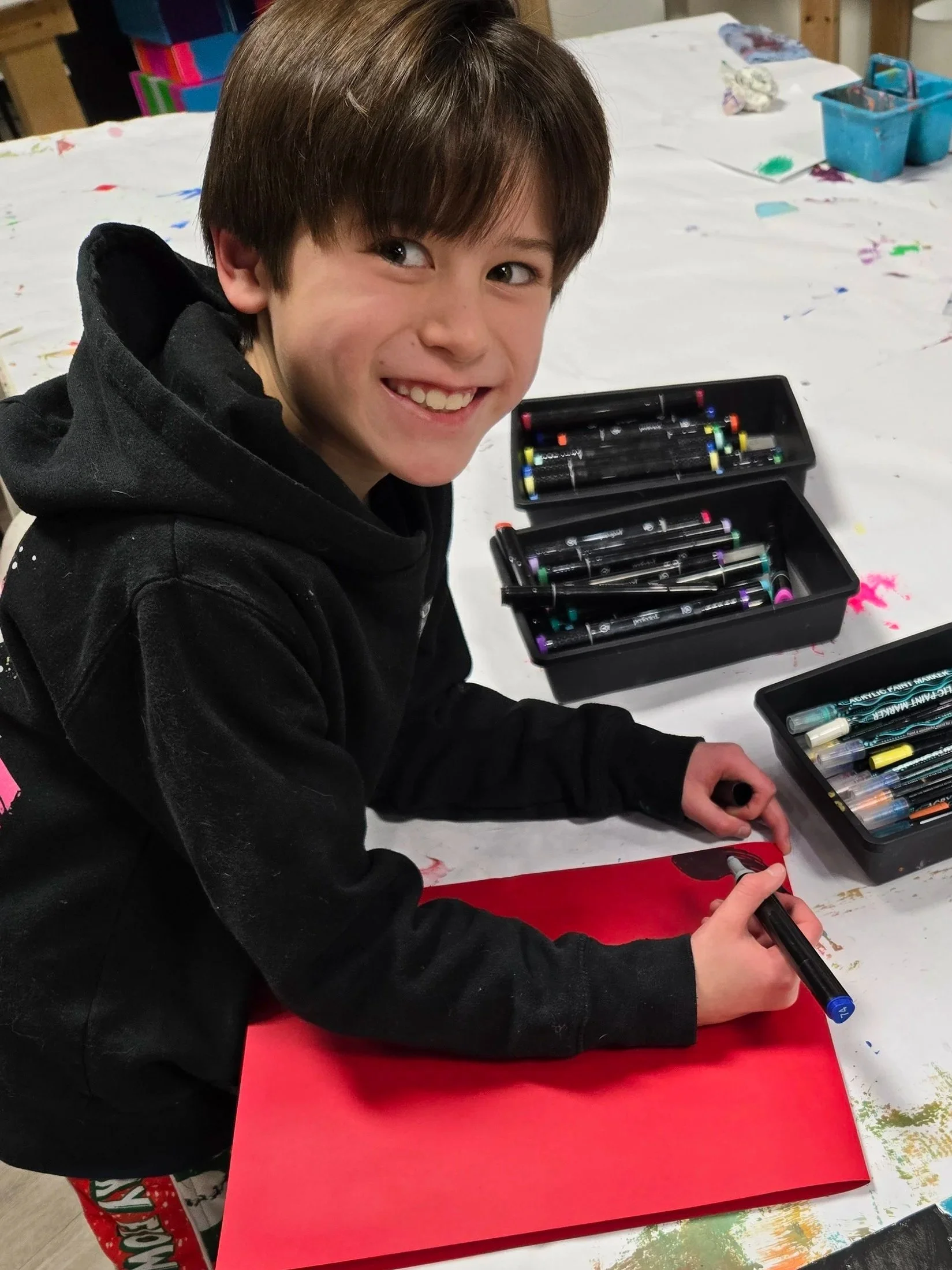 Young boy smiling at a table with art supplies, including markers and a colorful painted picture of a dog with a rainbow background.