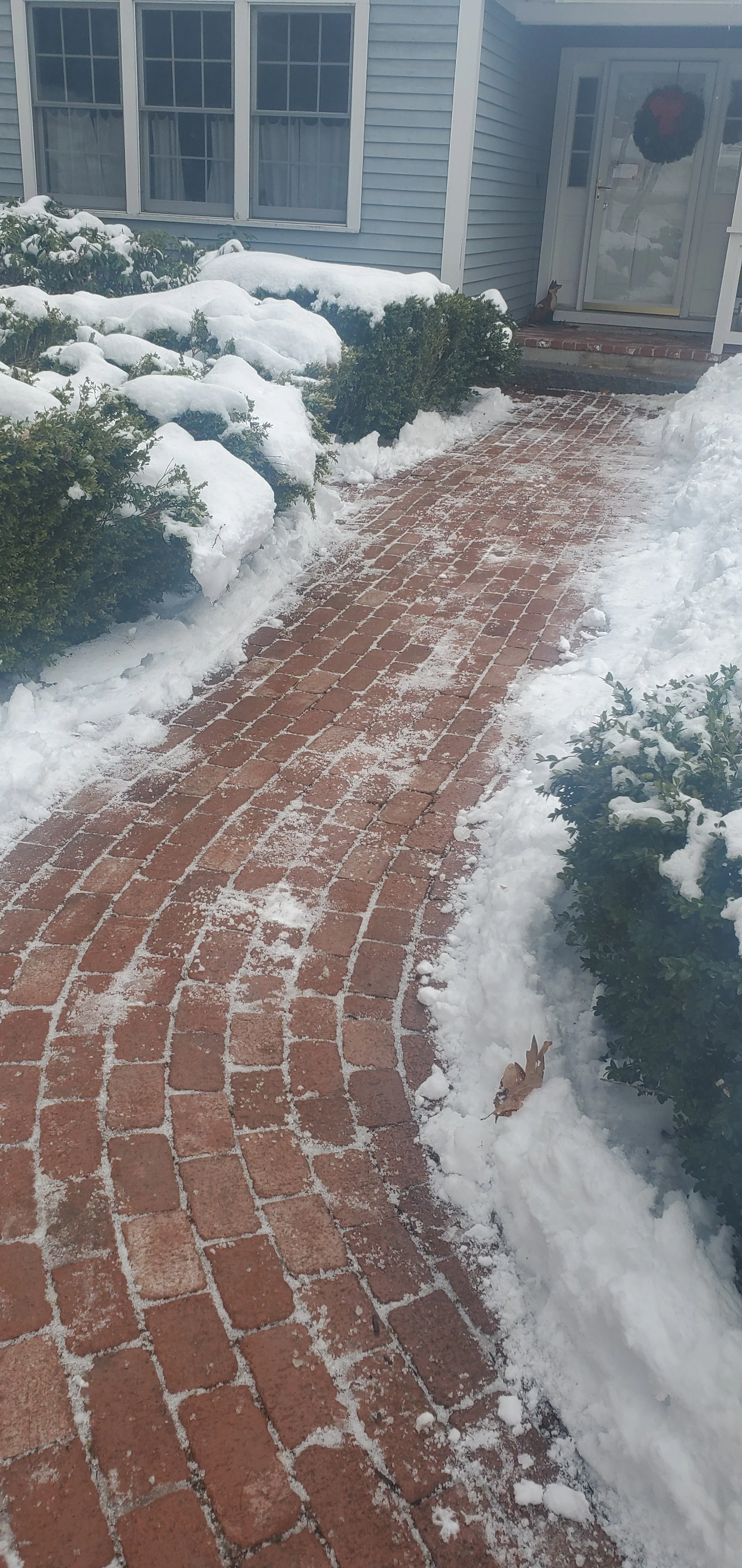 A snow-covered brick pathway leading to a house entrance. The path is lined with snow-covered bushes and the house has a light blue exterior. A wreath hangs on the front door.