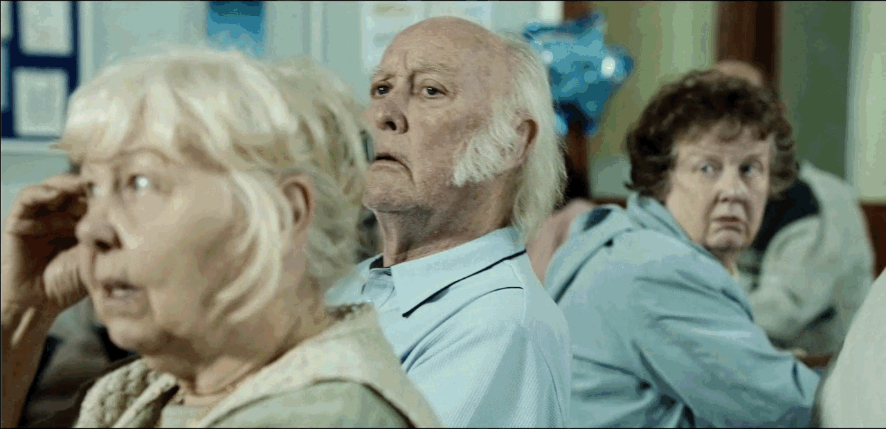 Three elderly people sitting on a bench, with one looking to the side and the others looking forward, in a room with a blue balloon in the background.