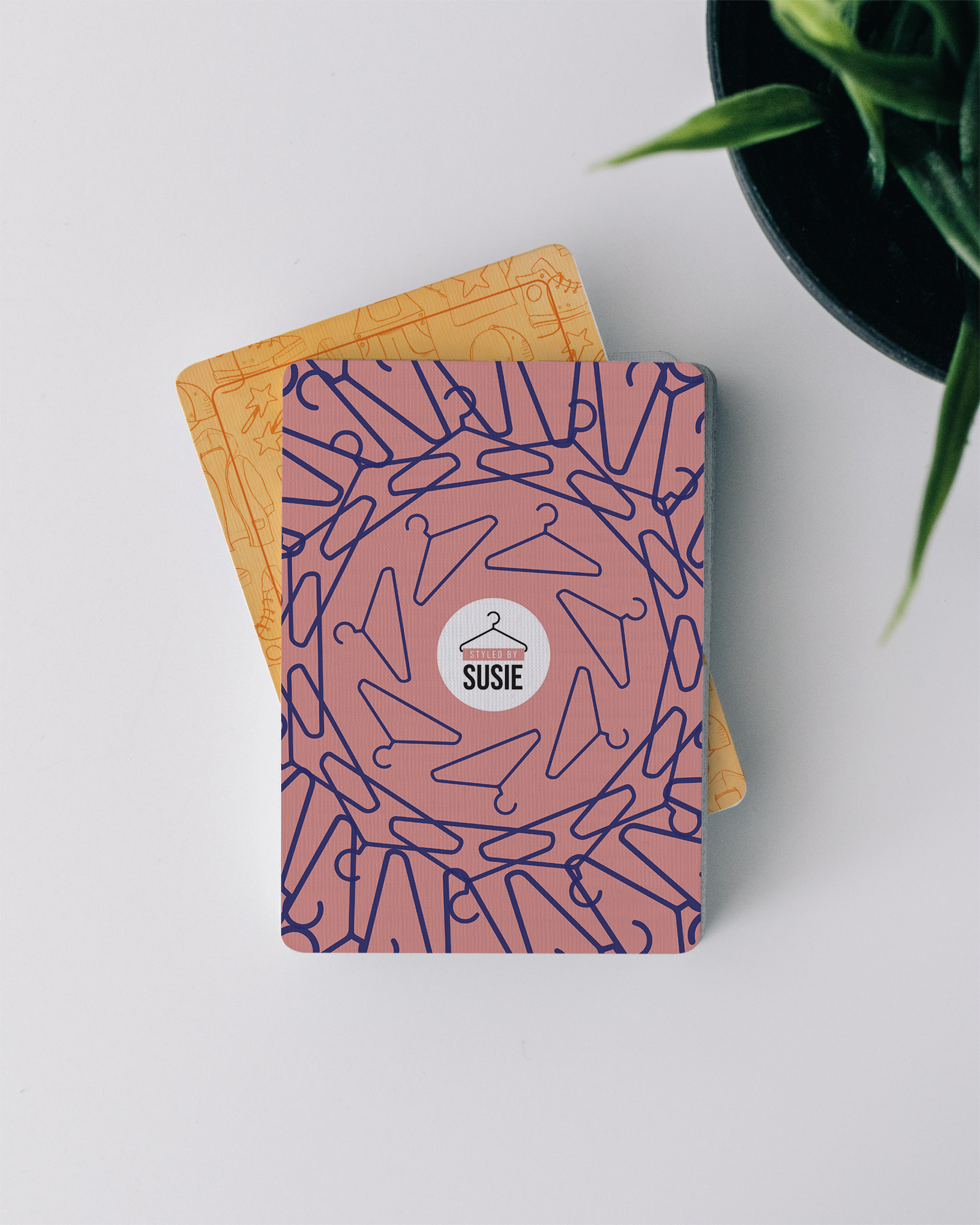 A pink card with a pattern of purple clothes hangers, featuring a circular sticker with a hanger illustration and the name 'Susie' on a white background, placed on top of a yellow card with a similar hanger pattern, next to a green plant in a black pot.