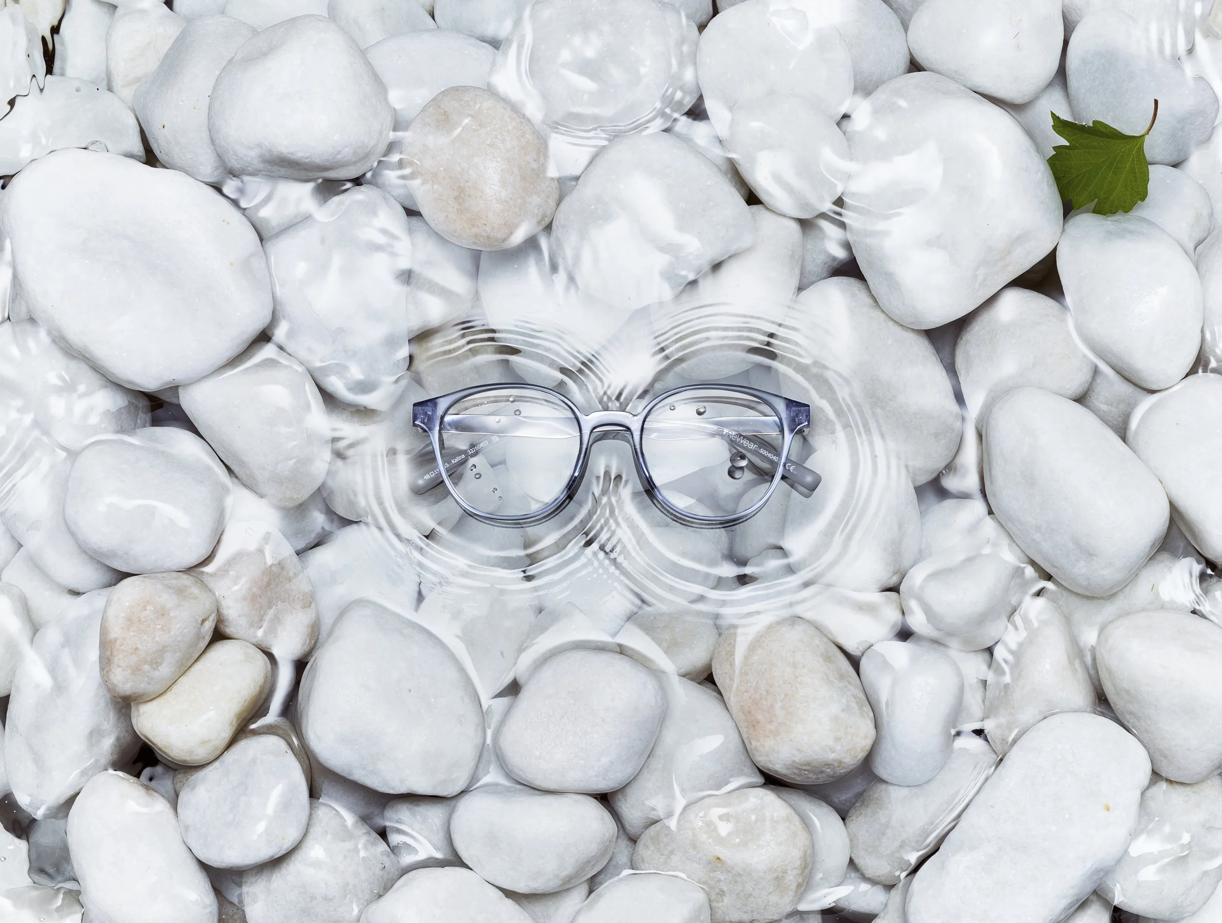 A pair of eyeglasses submerged in water, resting on white pebbles near a green leaf.