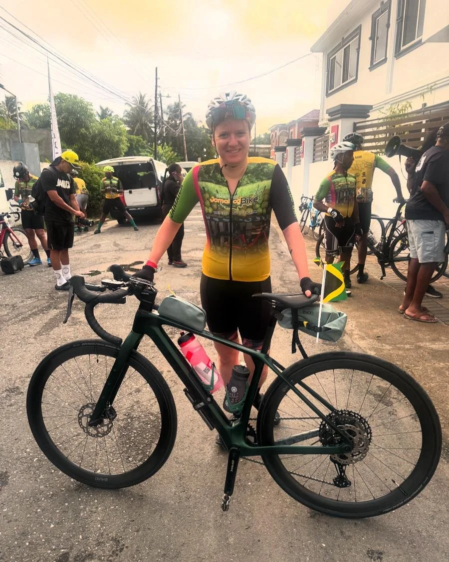 A woman wearing a cycling helmet and colorful cycling jersey standing next to a black road bike with a water bottle and small bag attached, participating in a group bike ride event with other cyclists in the background.