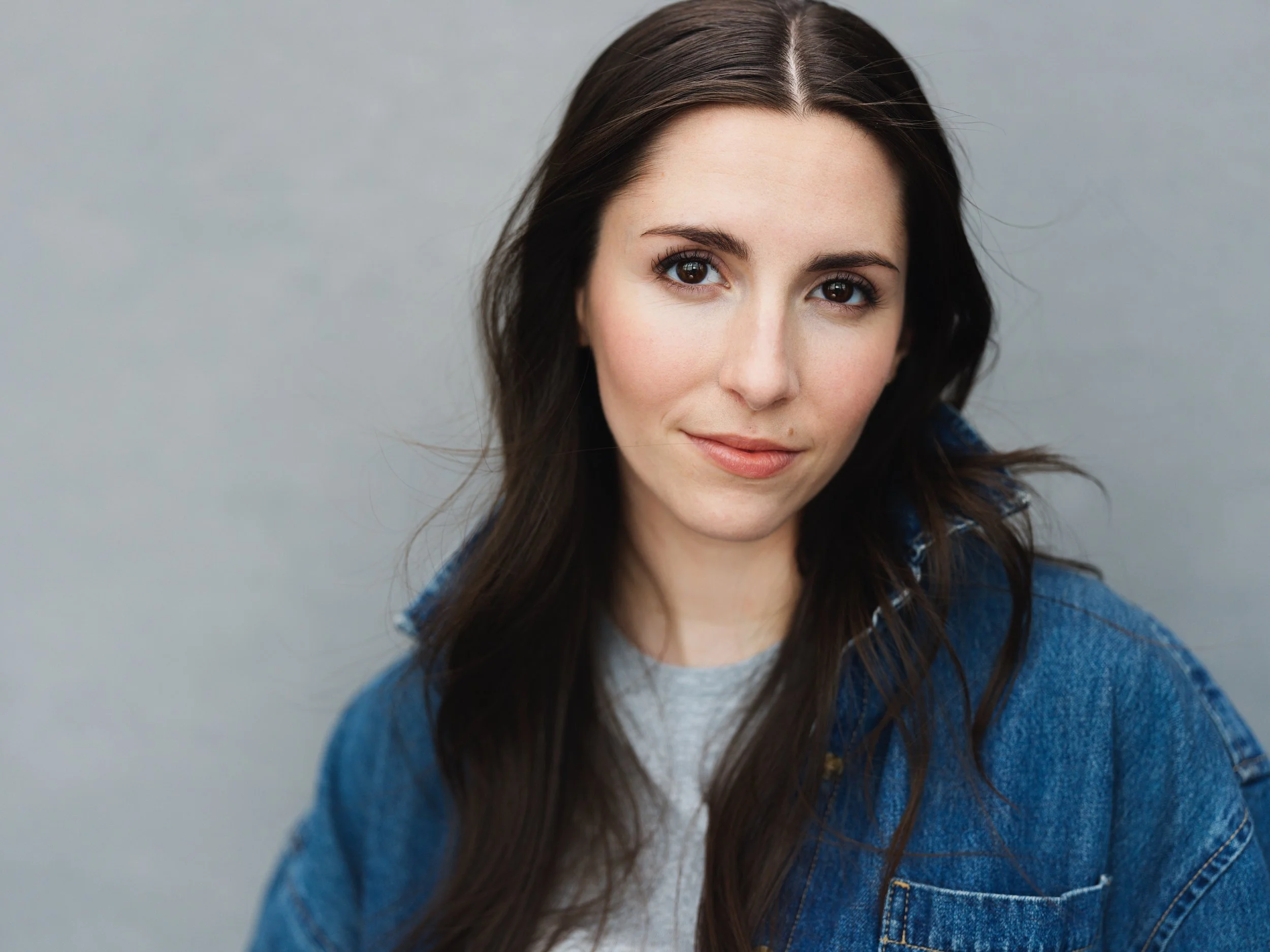 A woman with long dark brown hair, wearing a denim jacket and a green top, poses with arms crossed against a dark, textured background.
