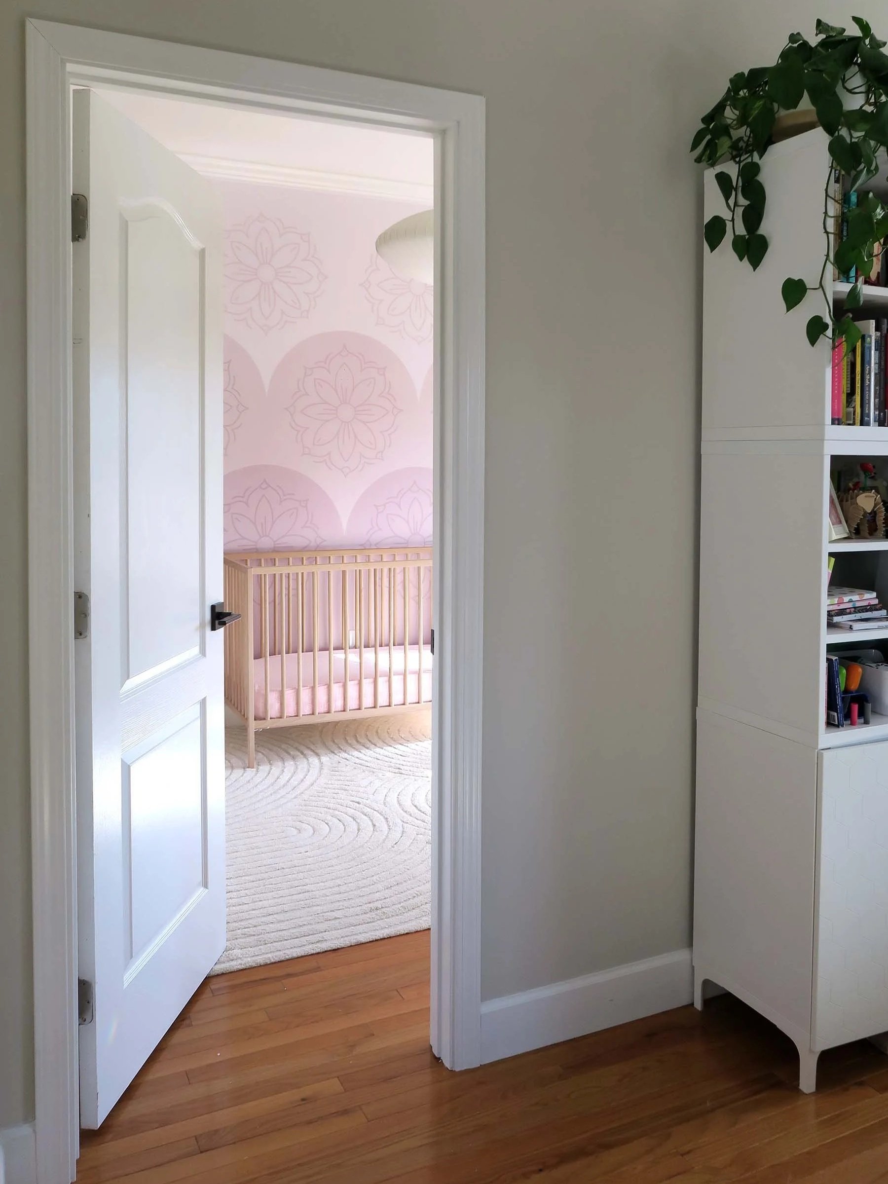 View of a doorway leading to a nursery with pink hand painted mural featuring large modern indian floral patterns, a wooden crib with pink bedding, and a white rug on a wooden floor. SF Bay Area mural artist interior design portfolio & inspiration.