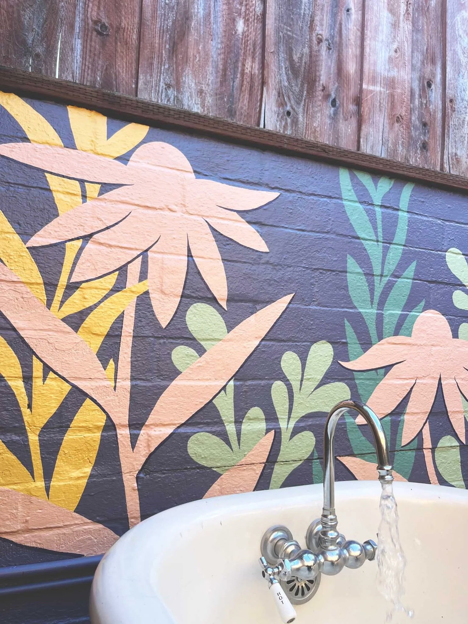 Faucet with running water against a colorful painted mural of various plants and flowers on a brick wall. SF Bay Area mural artist designer portfolio & inspiration.