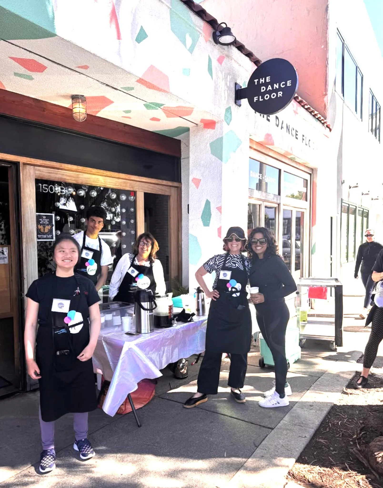 Group of people standing outside a storefront called 'The Dance Floor,' with some wearing aprons and event badges, and a table with drinks and snacks in front.