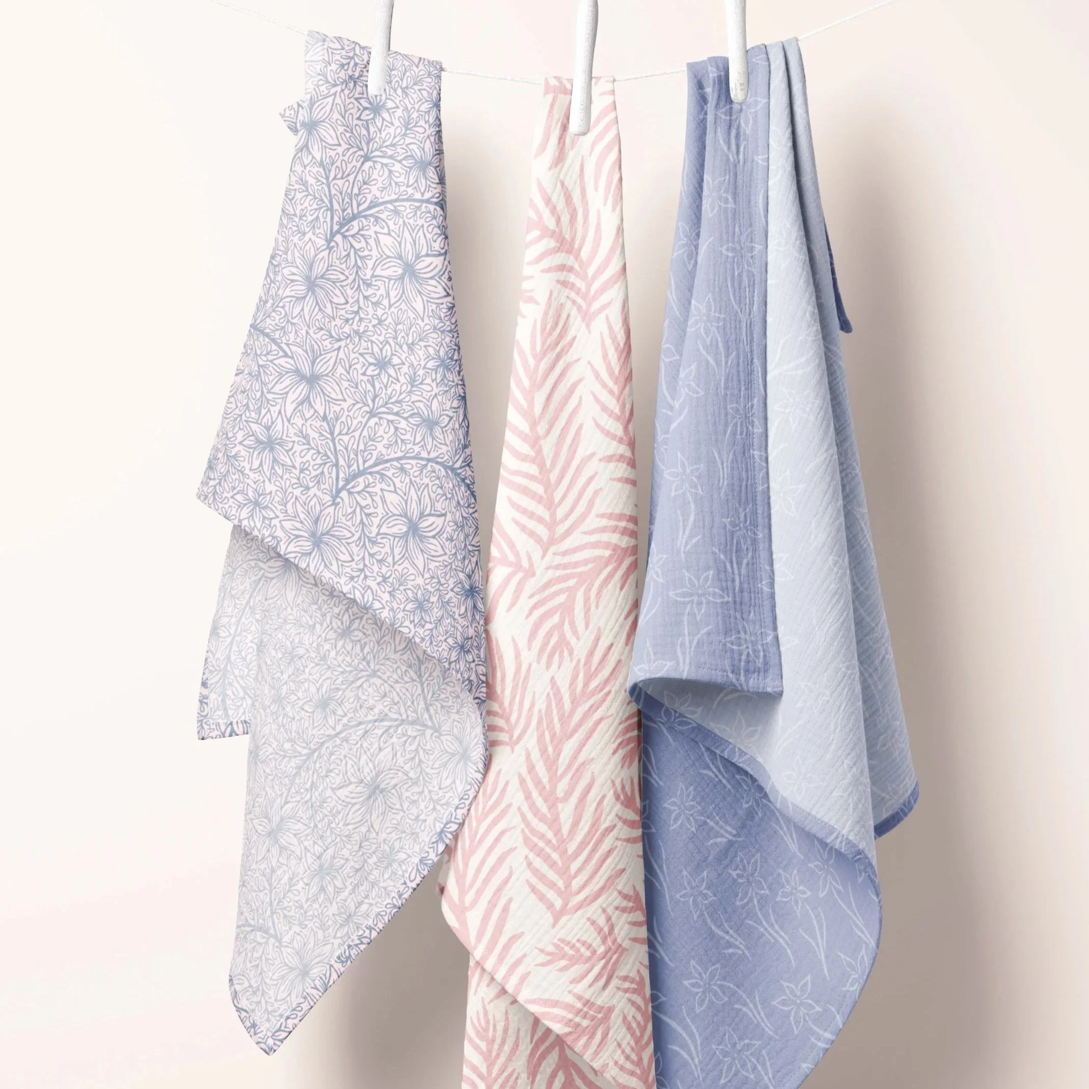Three pastel-colored towels with floral and leaf patterns hanging on a white clothesline against a plain white wall. SF Bay Area mural artist and illustrator surface pattern designer portfolio available for art licensing.