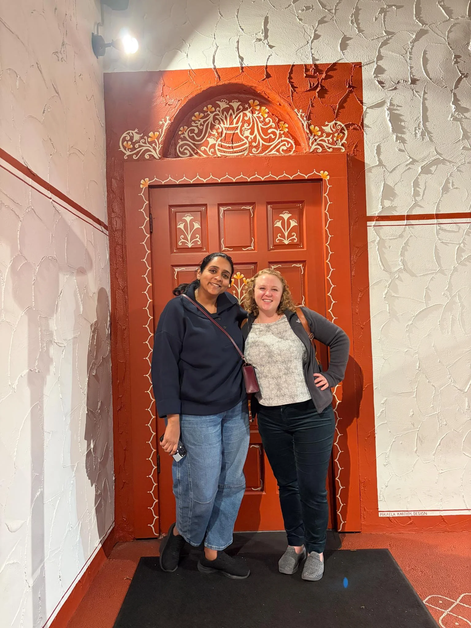 Two women standing in front of a decorative red door with ornate white and yellow floral patterns, inside a room with white textured walls.