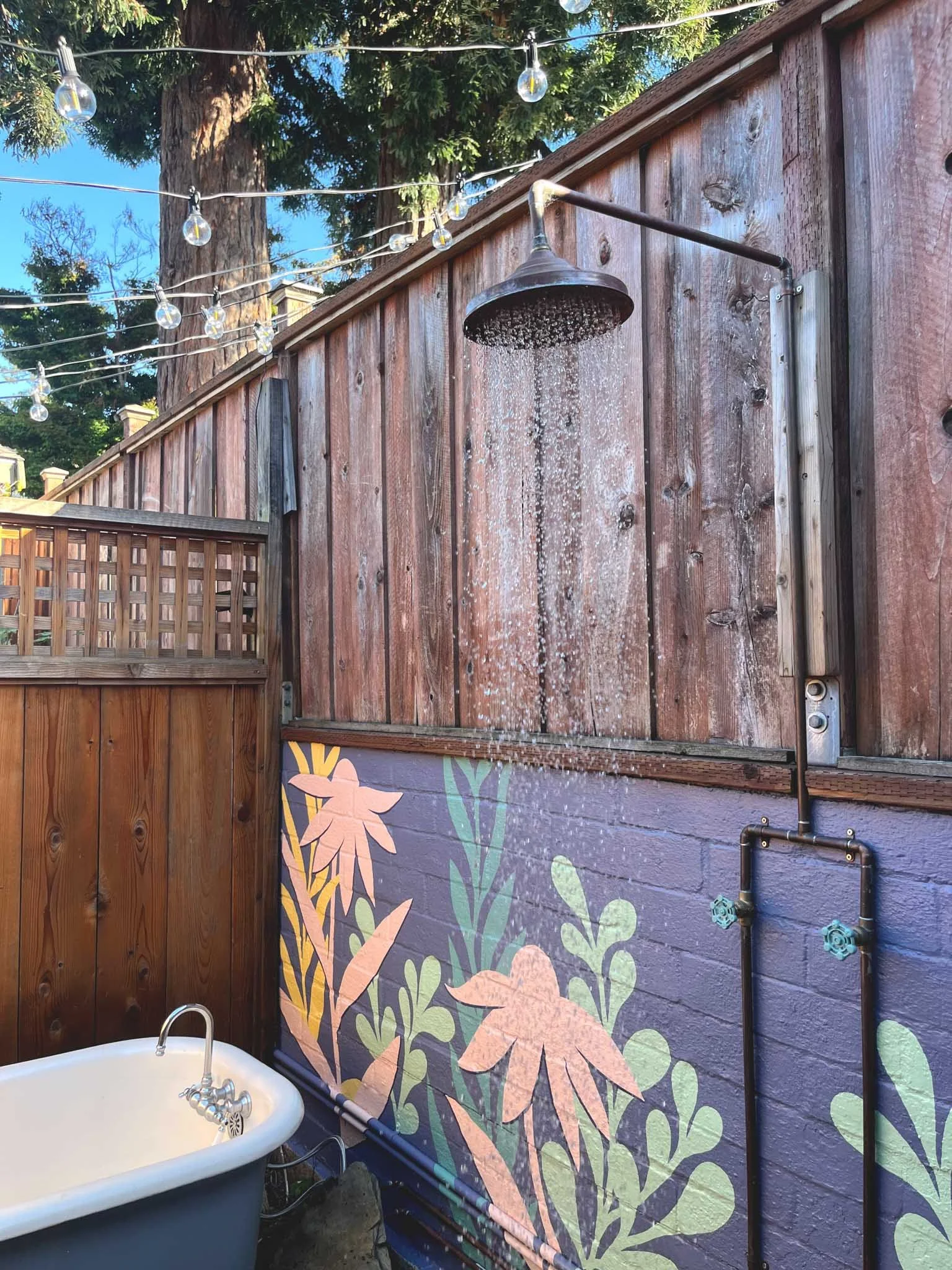 Outdoor outdoor shower mounted on a wooden fence with colorful flower mural, a bathtub below, string lights overhead, and trees in the background. SF Bay Area mural artist designer portfolio & inspiration.