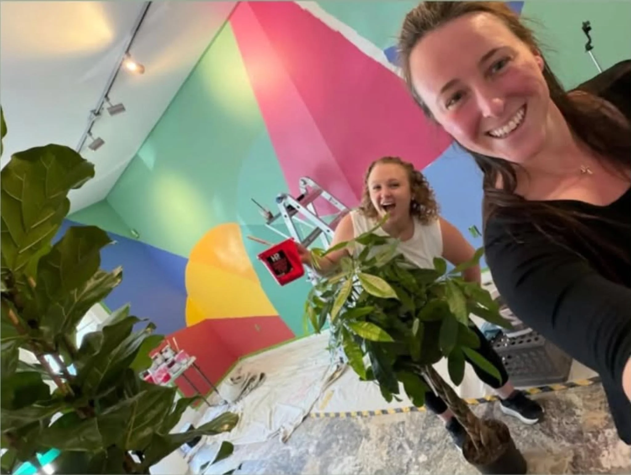 Smiling young women taking a selfie in front of a colorful mural with a ferris wheel, with greenery in the foreground.