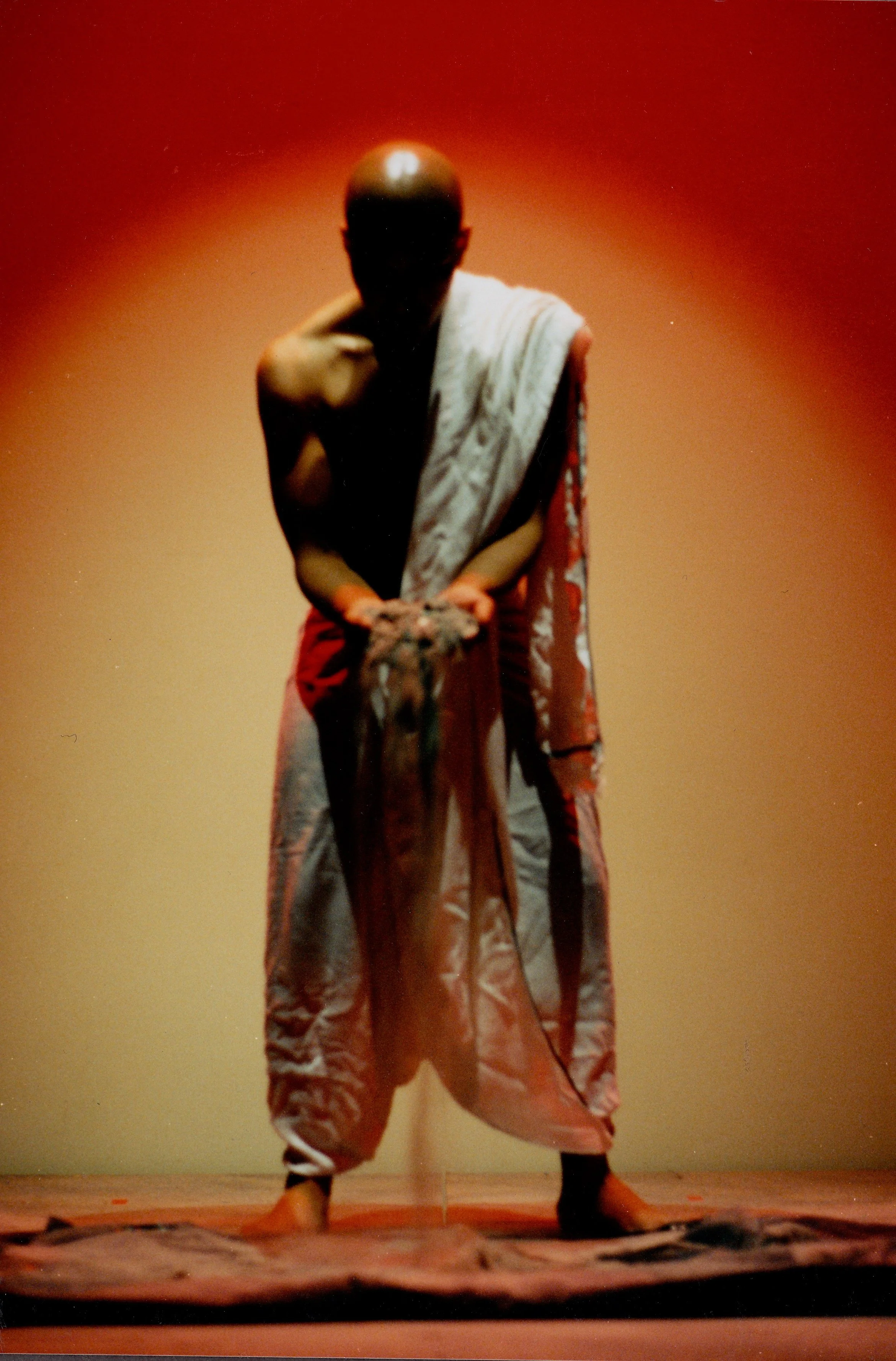 A man performing a traditional dance on a stage, wearing loose-hipped pants and a towel over his shoulder against a warm background.