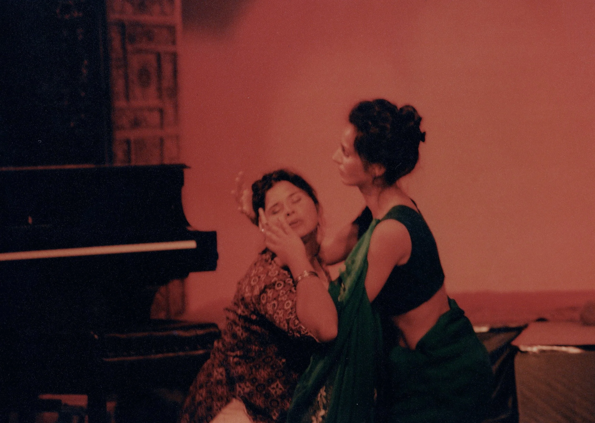 A woman with curly hair and glasses is dancing with another woman with short dark hair, wearing a green dress, in a dimly lit room with a red-orange background and a black upright piano.