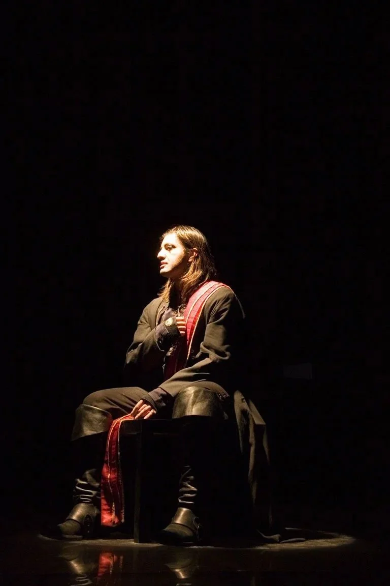 A woman with long dark hair, dressed in a black coat and boots, sitting on a black chair on a dark stage with spotlight.