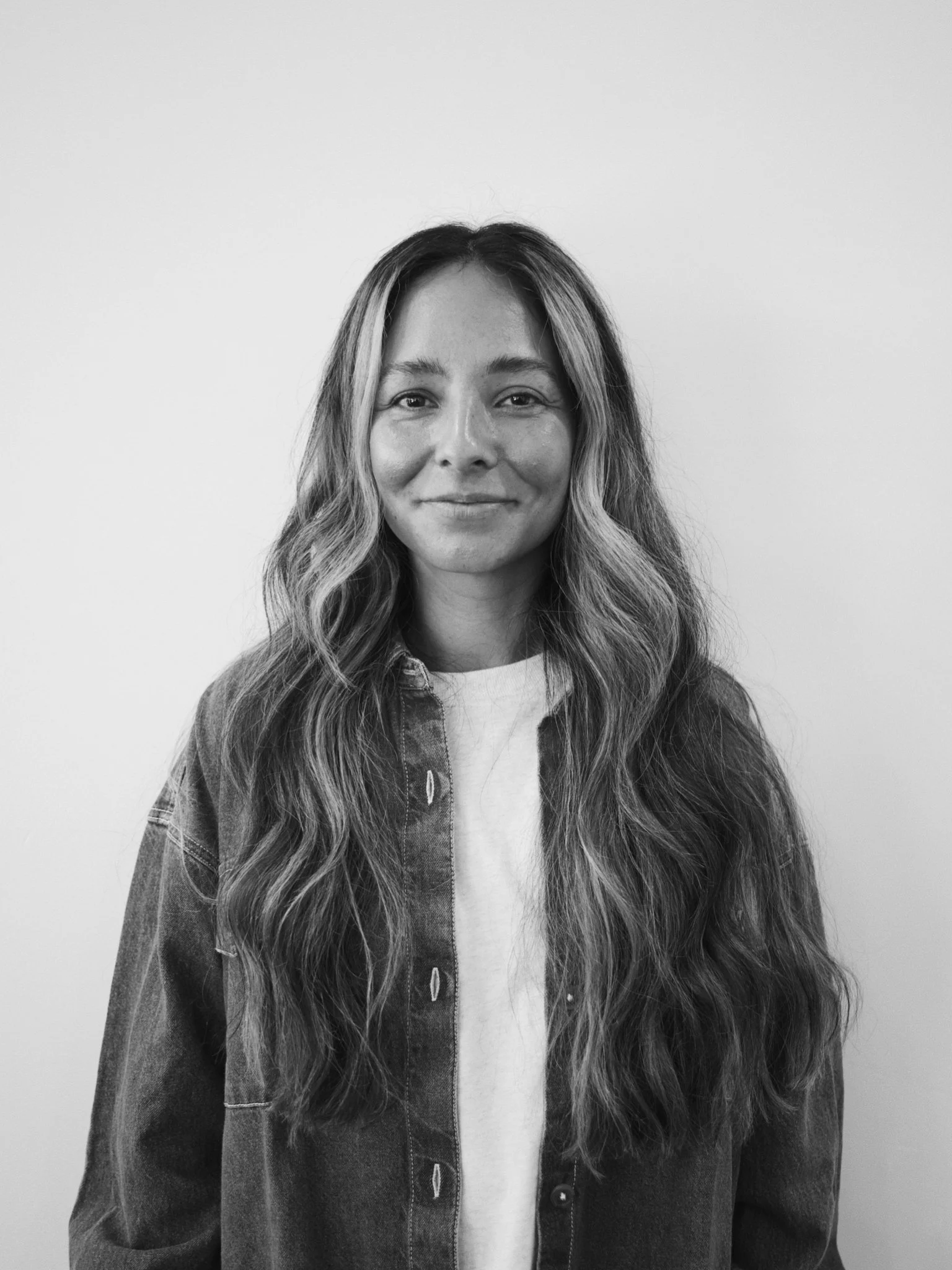Black and white portrait of a woman with long wavy hair, wearing a denim jacket over a white t-shirt, standing against a plain wall.