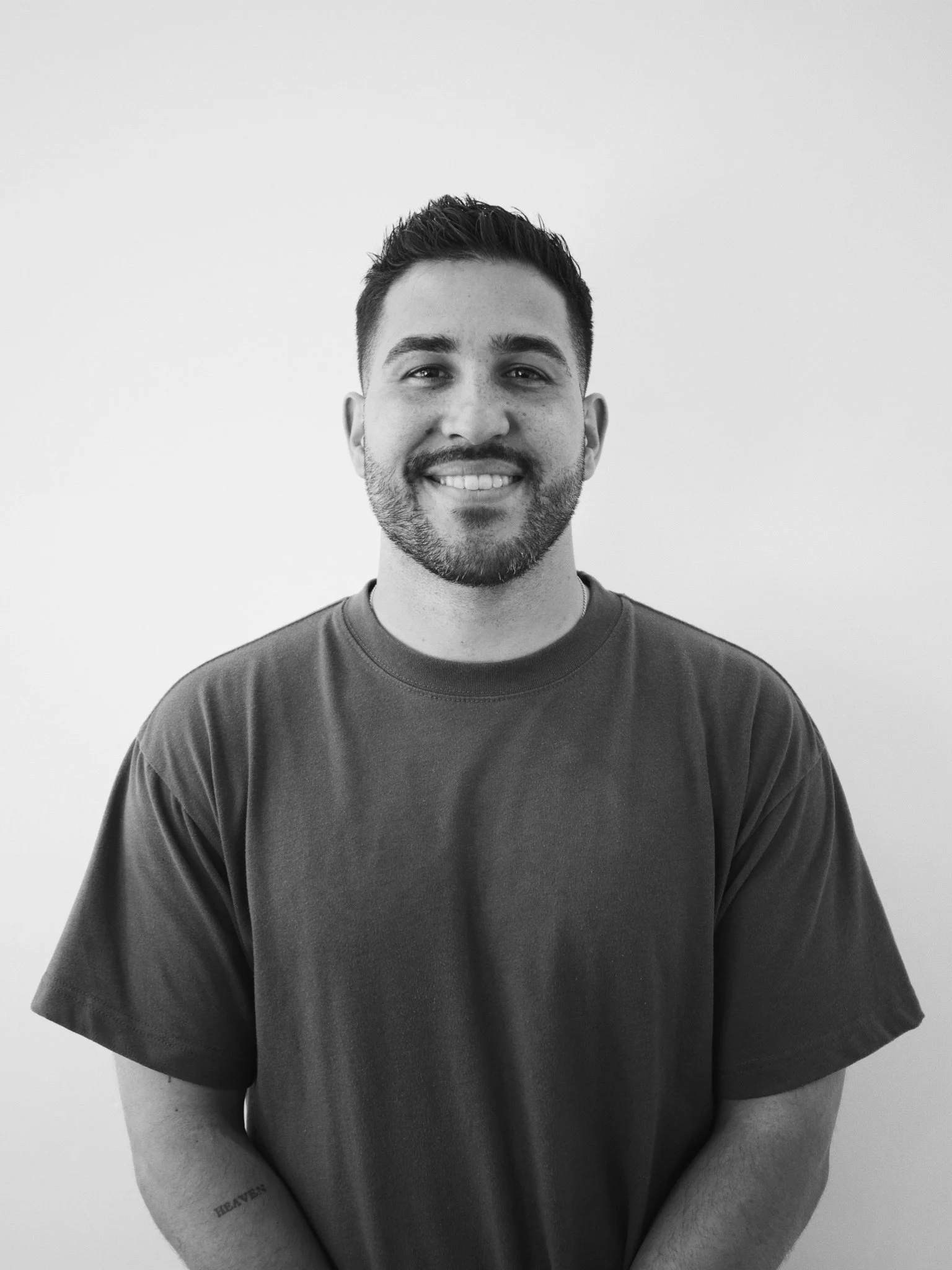 A young man with dark hair and a beard, smiling, wearing a plain dark T-shirt, standing against a plain light-colored wall.