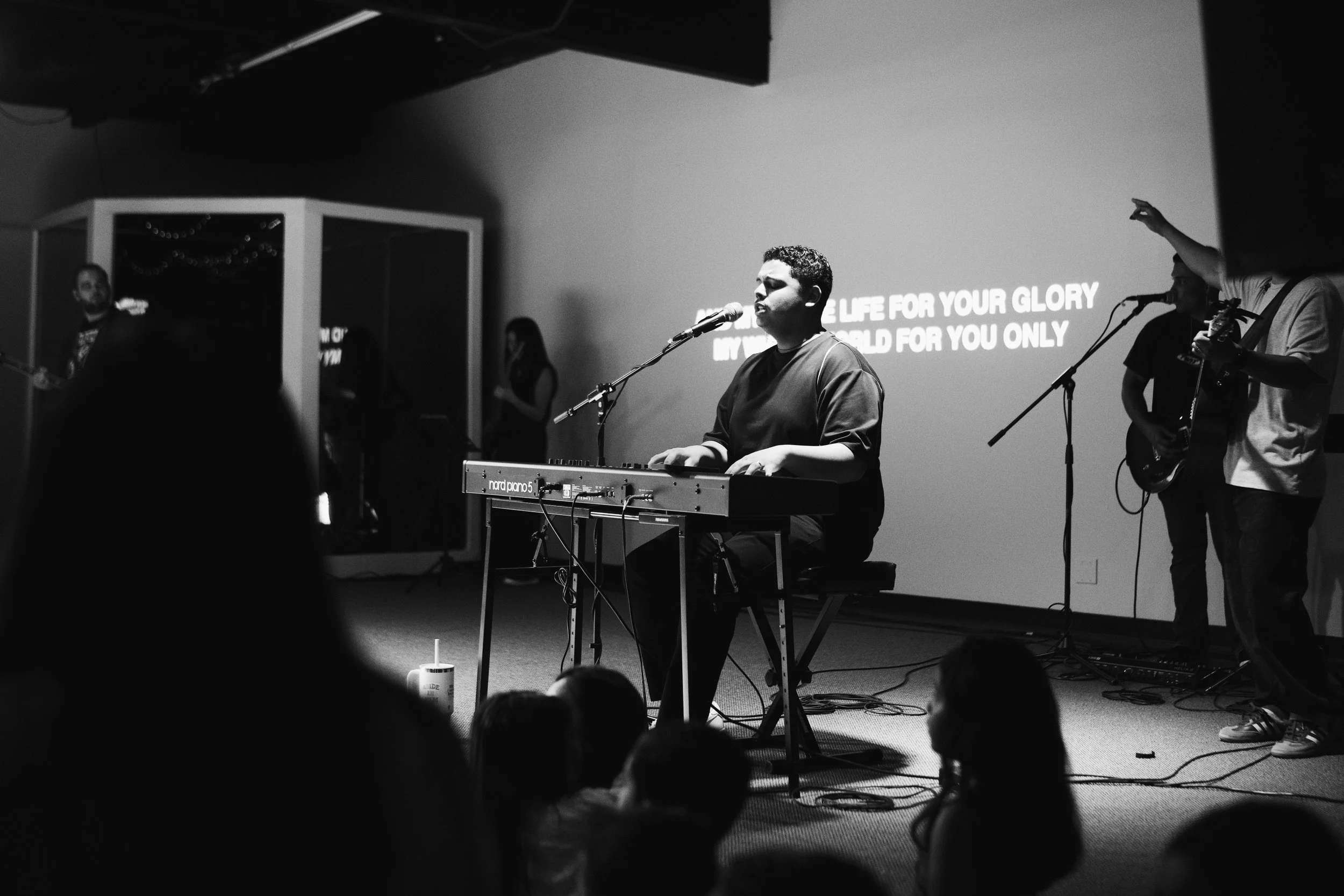 A black and white photo of a musical performance in a church-like setting, with a man playing a keyboard and singing into a microphone, surrounded by other musicians and singers, with lyrics projected on the wall behind them.