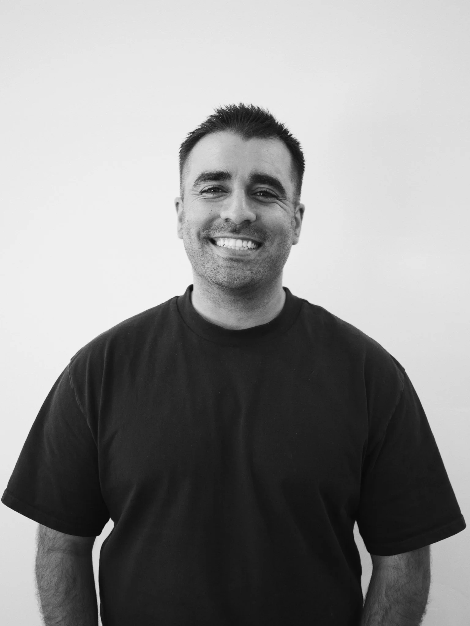 A smiling man with short dark hair, wearing a plain black t-shirt, stands against a light-colored plain background.