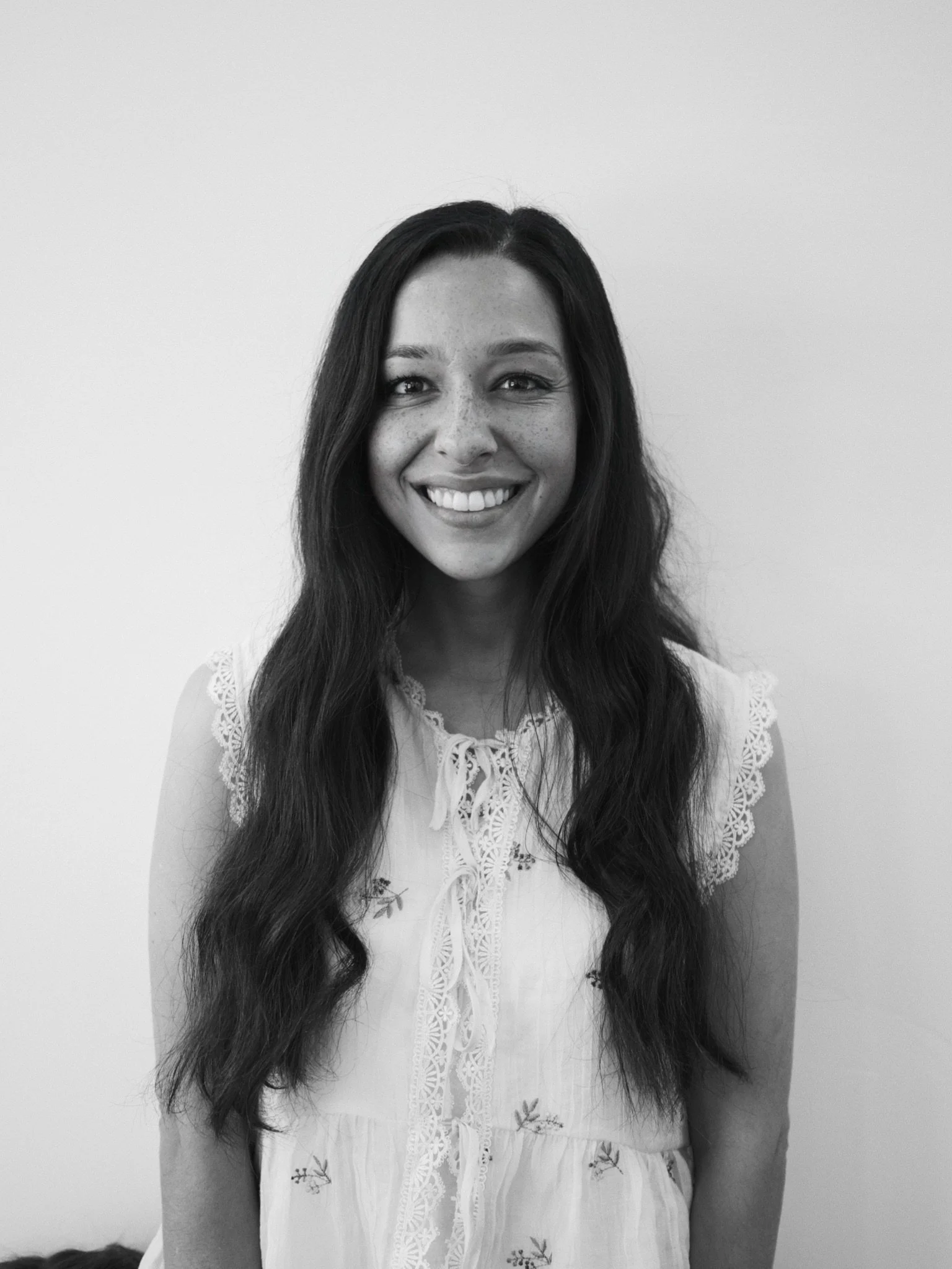 Black and white photo of a smiling woman with long wavy hair, wearing a light-colored top with embroidered floral details and lace accents on the sleeves.