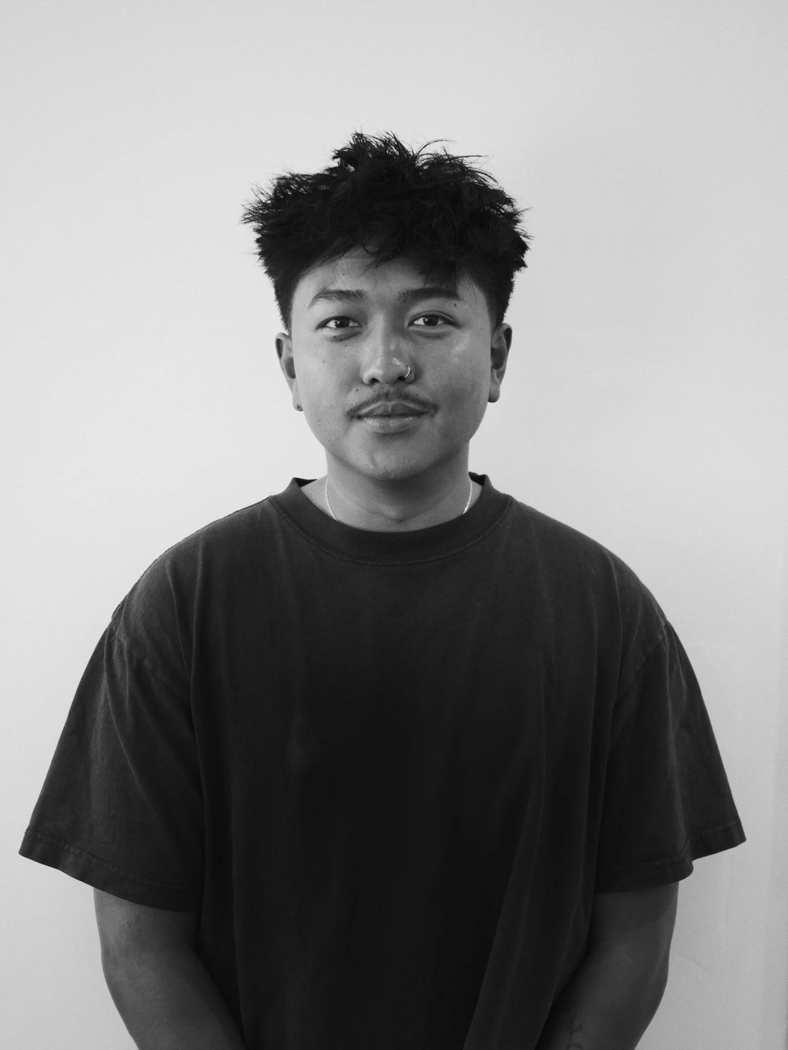 Black and white portrait of a young person with short, messy hair, wearing a black t-shirt, standing against a plain light-colored background.