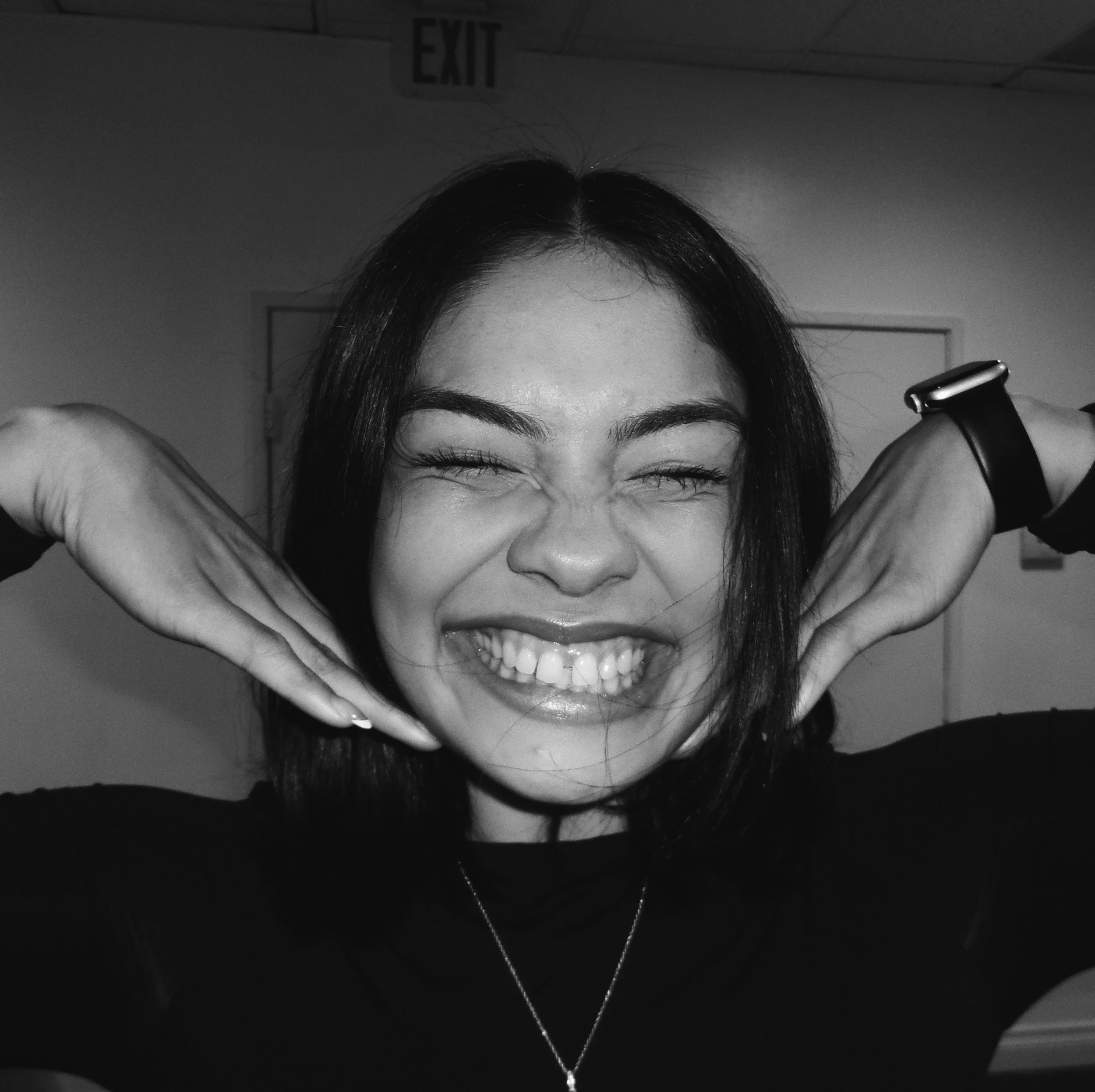 Woman with dark hair posing with a big smile, eyes closed, hands framing her face, in black and white