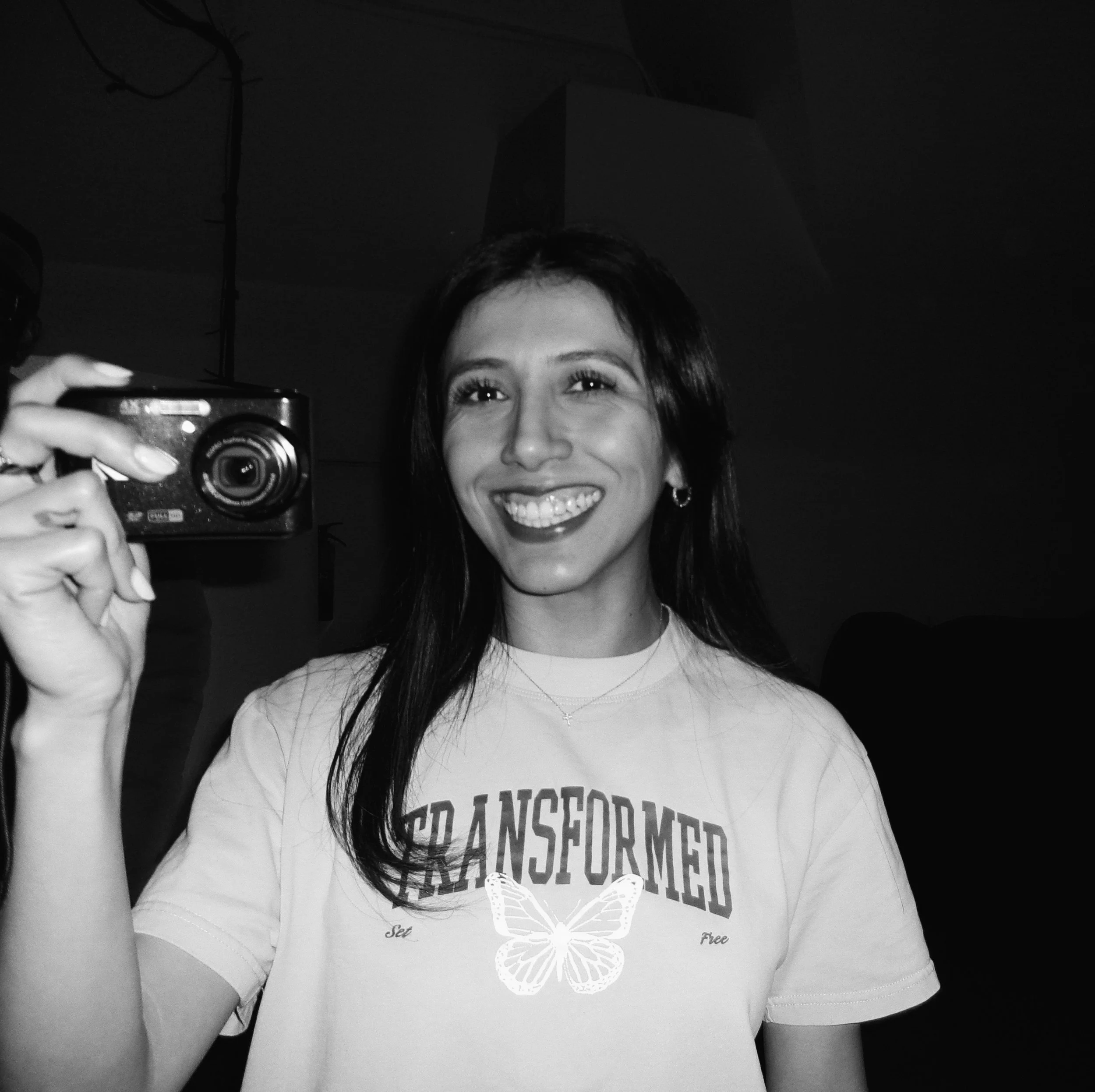 A smiling woman with long dark hair, wearing a T-shirt with butterfly and text, holding a camera, in a dark indoor setting.