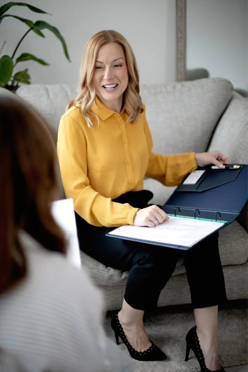 A woman with blonde hair, wearing a yellow blouse and black pants, is sitting on a gray sofa. She appears to be talking or explaining something to another person who is partially in the foreground. She is holding a binder or planner with papers inside, and there are folders on her lap. The setting looks like a modern living room with a green plant in the background.