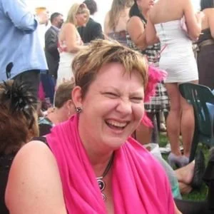 Woman with short hair laughing, wearing a pink scarf, at a crowd of people outdoors.