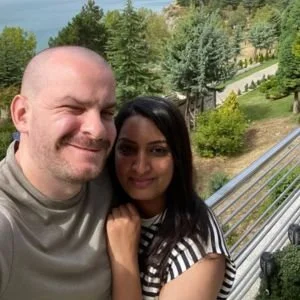 A man and a woman smiling outdoors on a sunny day, with greenery and trees in the background.