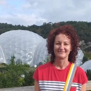 A woman with curly red hair wearing a red and white striped shirt, standing outdoors with large geodesic domes in the background and greenery around.