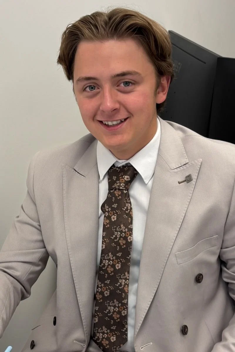 A young man with brown hair and a friendly smile, dressed in a light beige suit with a white shirt and a dark floral tie, sitting in an office with a plain light-colored wall and a black monitor in the background.