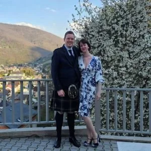 A man and woman standing on a balcony with a scenic view of mountains and a river, smiling at the camera. The man is dressed in a kilt and suit, the woman in a floral dress, with blooming trees nearby.