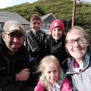 A family of five smiling outdoors, with a man, a woman, three children, and a house with trees in the background.