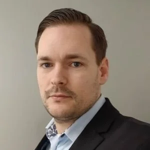 A man with short brown hair and a serious expression, wearing a suit and light blue shirt, against a plain background.