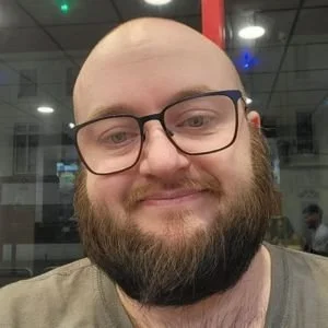 Close-up of a smiling man with glasses, a beard, and a shaved head, in an indoor setting with reflections of lights and people in the background.