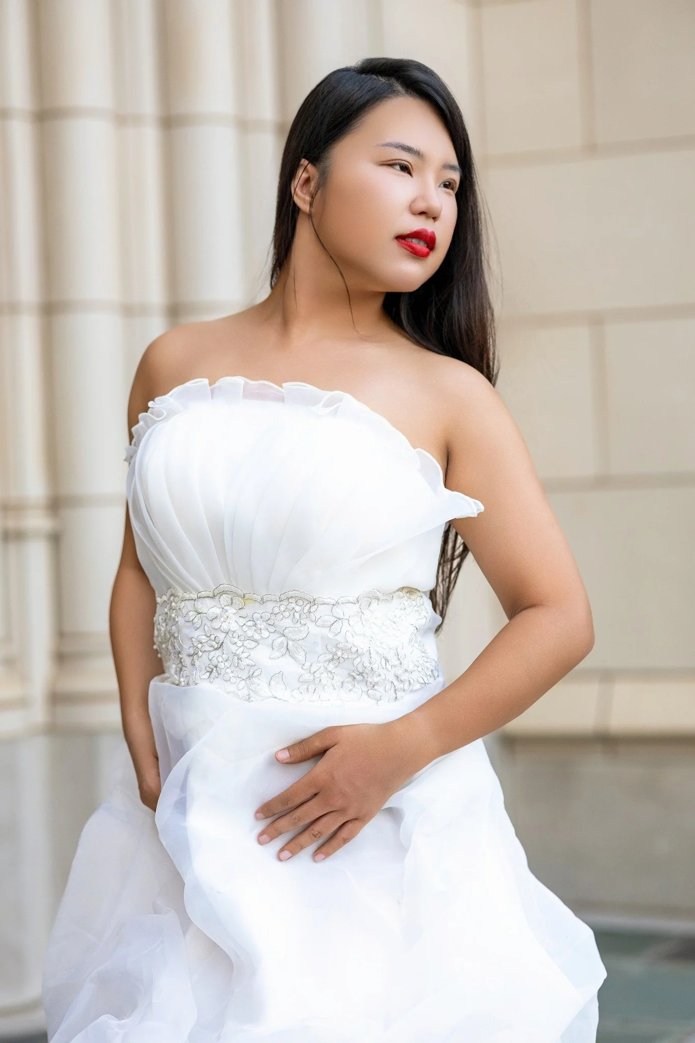 A woman in a strapless white dress with embroidery stands outdoors, looking to her right with long dark hair and red lipstick.