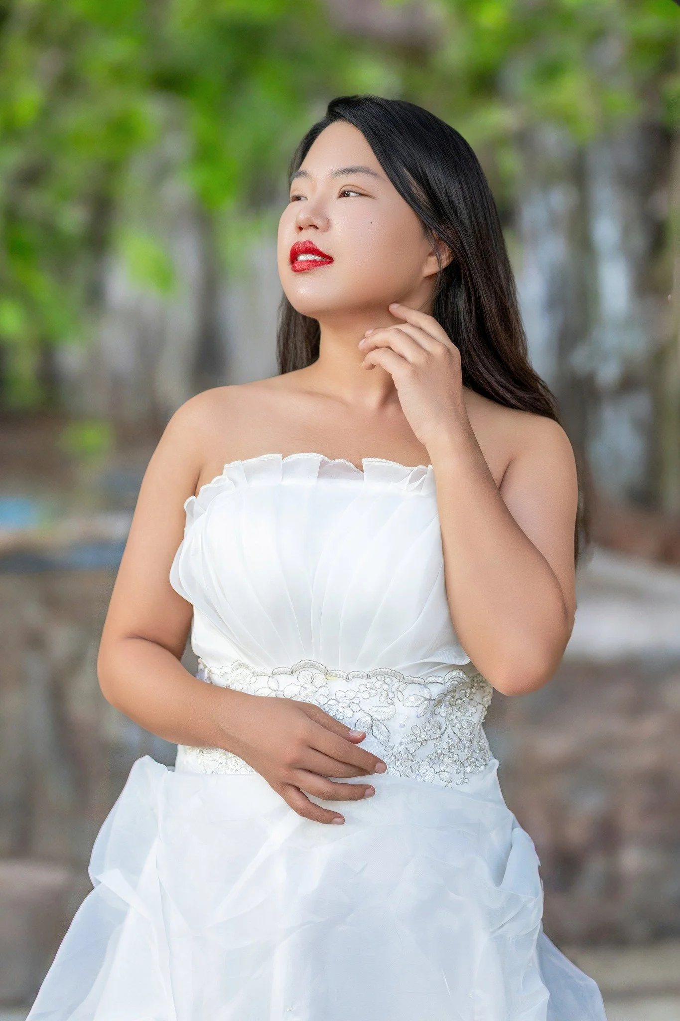 A woman wearing a white strapless dress standing outdoors with green trees in the background.