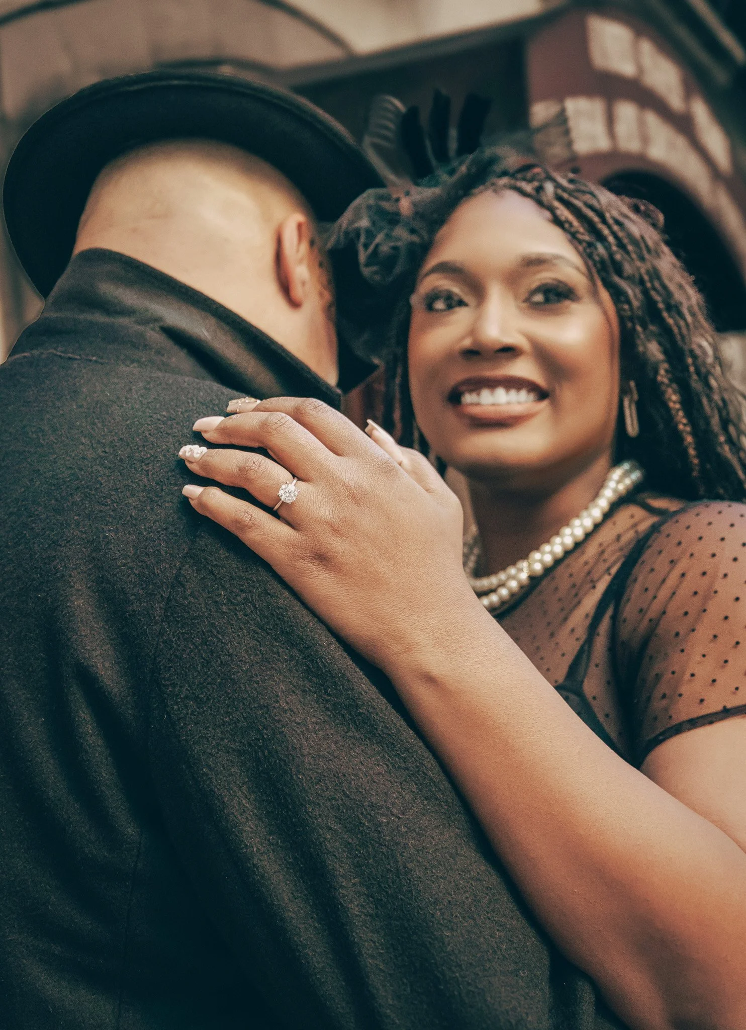A woman smiling and wearing a pearl necklace and earrings, holding a man close who is wearing a black hat and coat. She has her hand on his shoulder, showing a diamond ring.