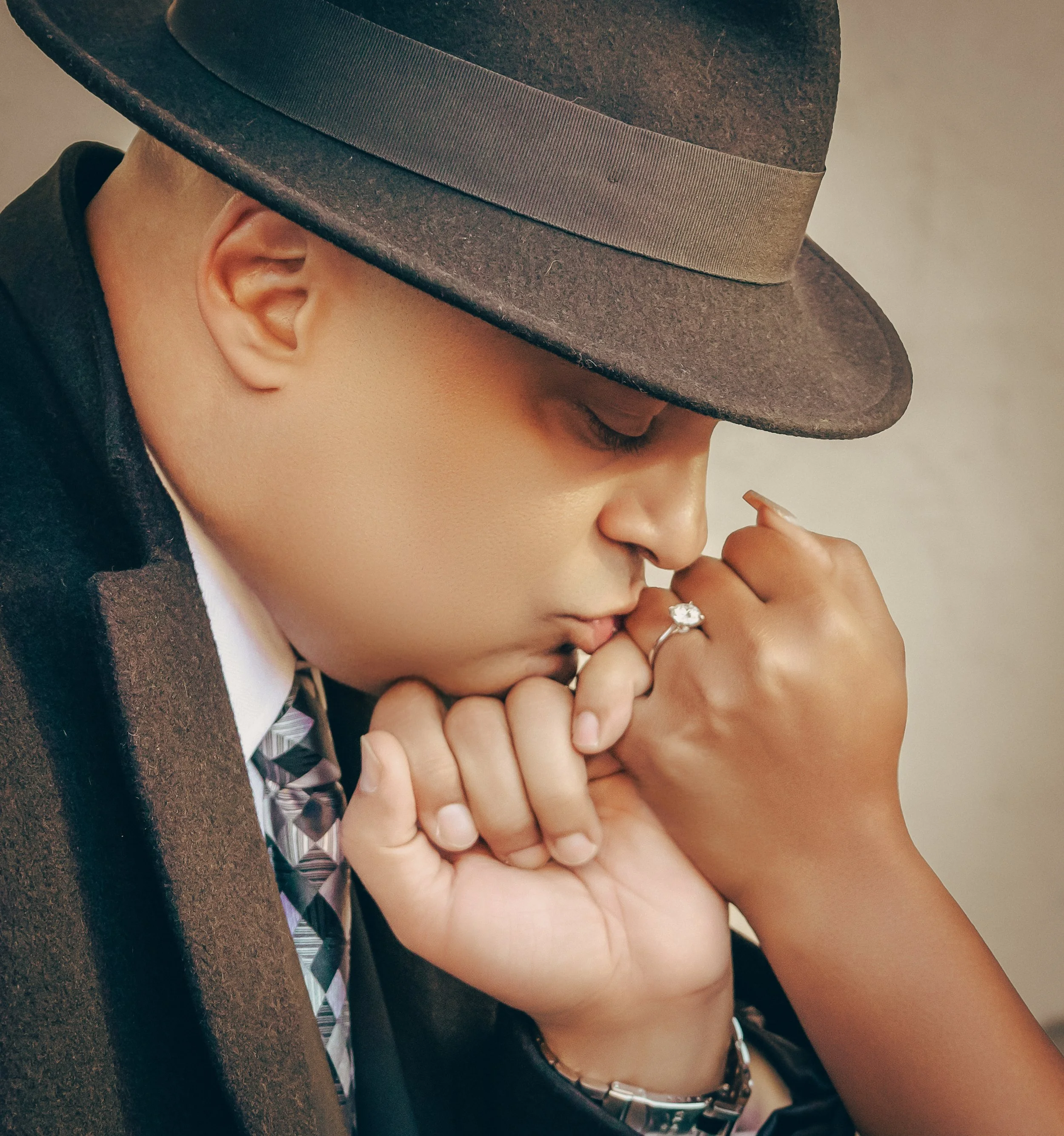 A person wearing a brown fedora hat, dressed in formal attire, with their eyes closed, gently holding and kissing a hand with a ring on the finger.