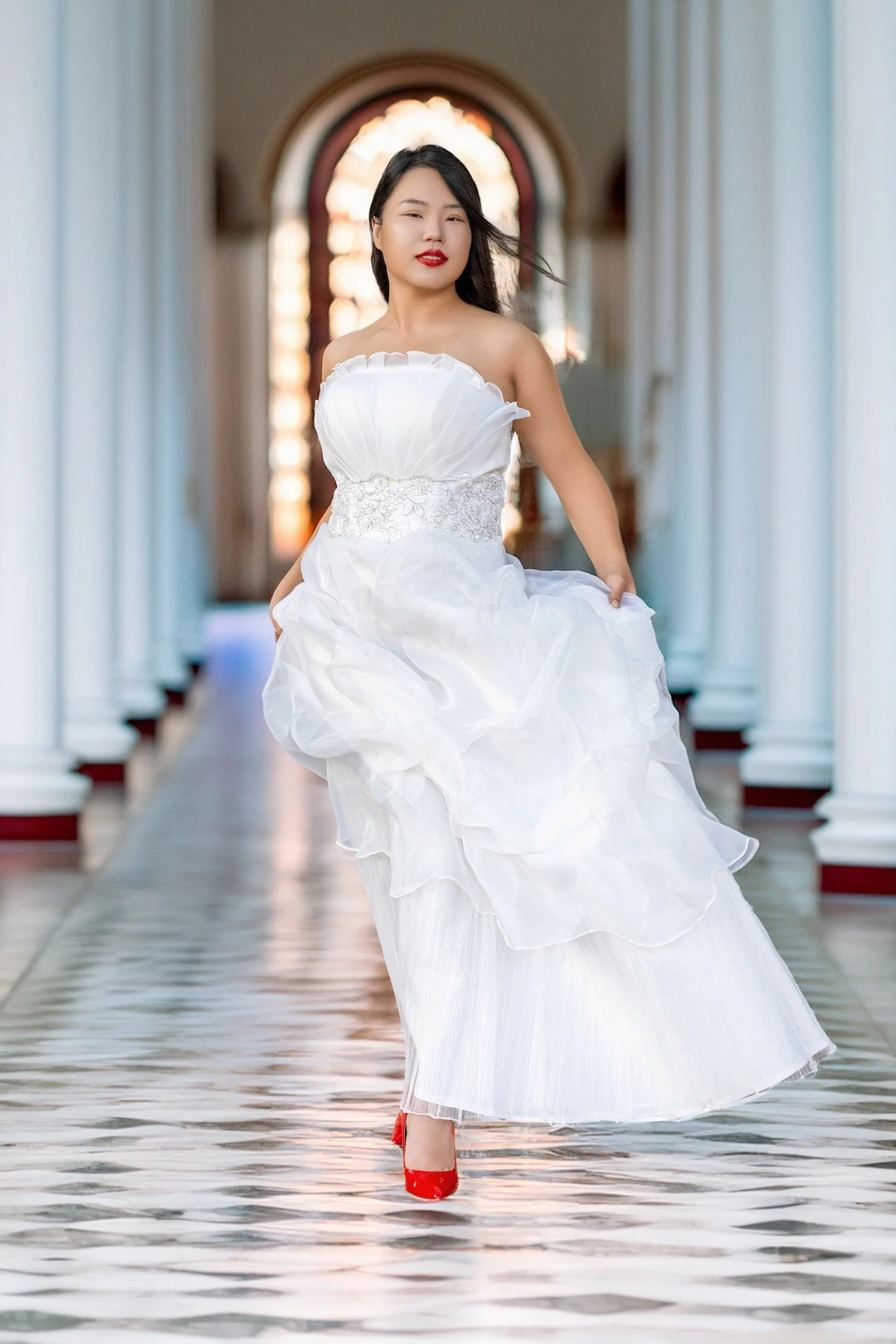 Woman in a strapless white wedding dress walking down a corridor with white columns and a patterned floor, holding the skirt with one hand, and wearing red shoes.