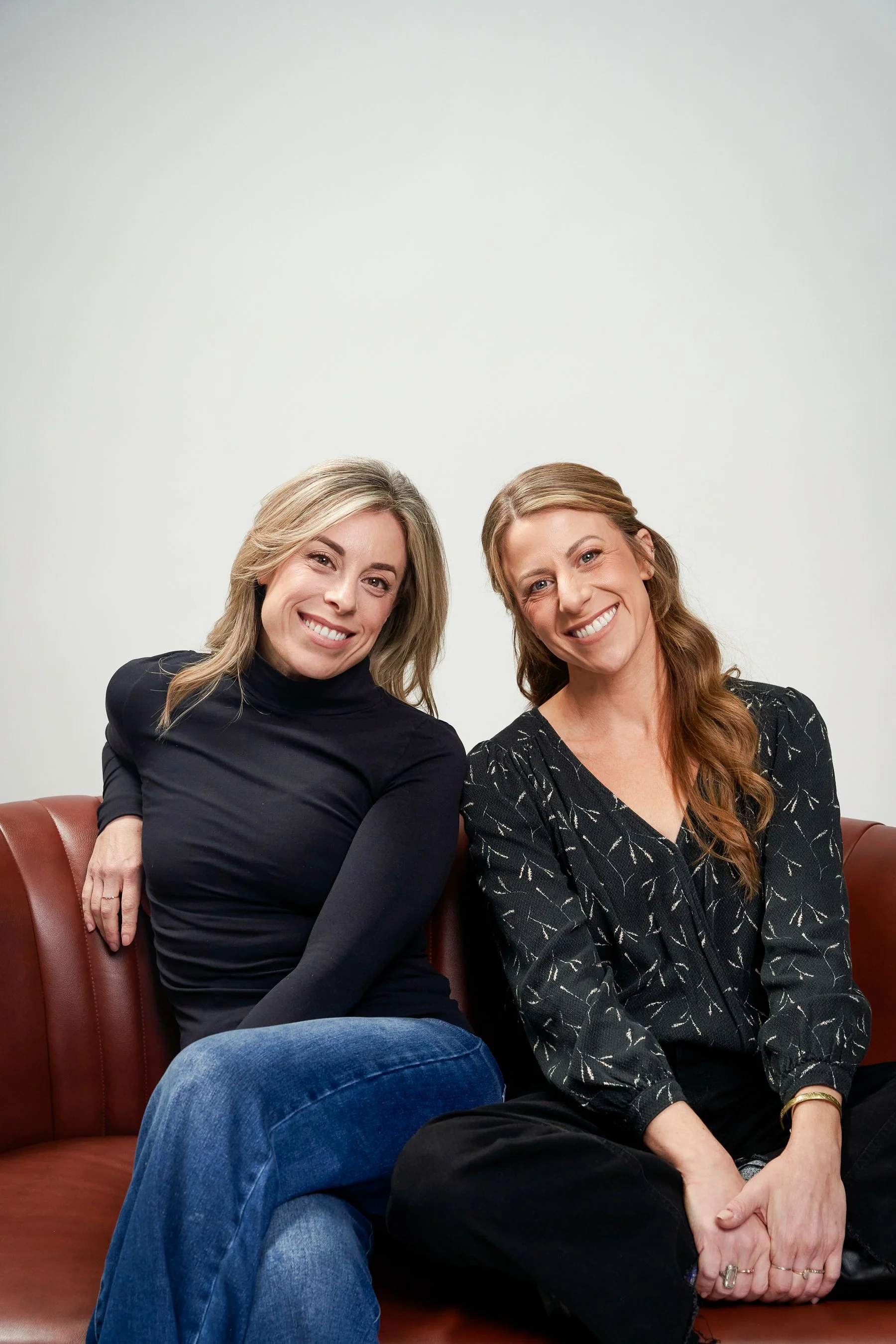 Two women sitting on a brown leather sofa, smiling at the camera, against a plain white wall.