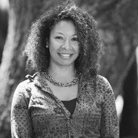 A woman smiling outdoors with curly hair, wearing a patterned blouse and necklace.