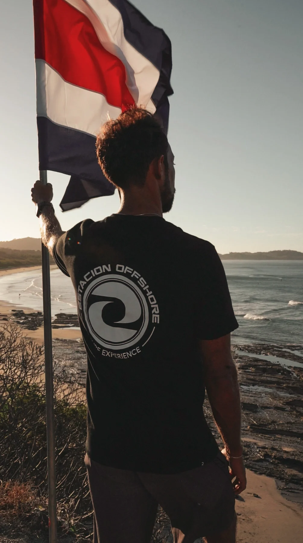 Surf instructor standing on Playa Guiones Nosara Costa Rica at sunset, holding a flag with the coastline and ocean in the background.