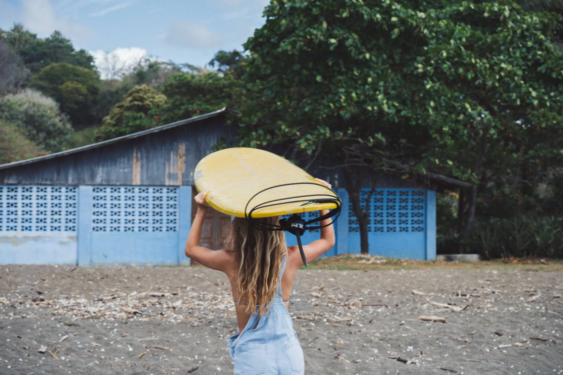 A surfer girl carrying a surfboard on the beach at Playa Ostional during a surf trip in Costa Rica, lush green trees in the background.