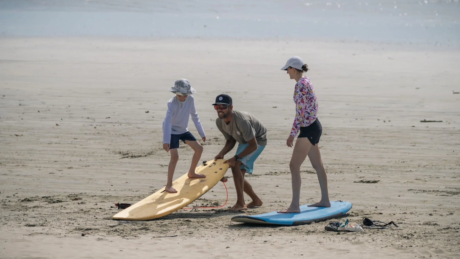 Family surf lesson on the beach in Nosara Costa Rica, coach helping kid onto a surfboard with kid's mom nearby.