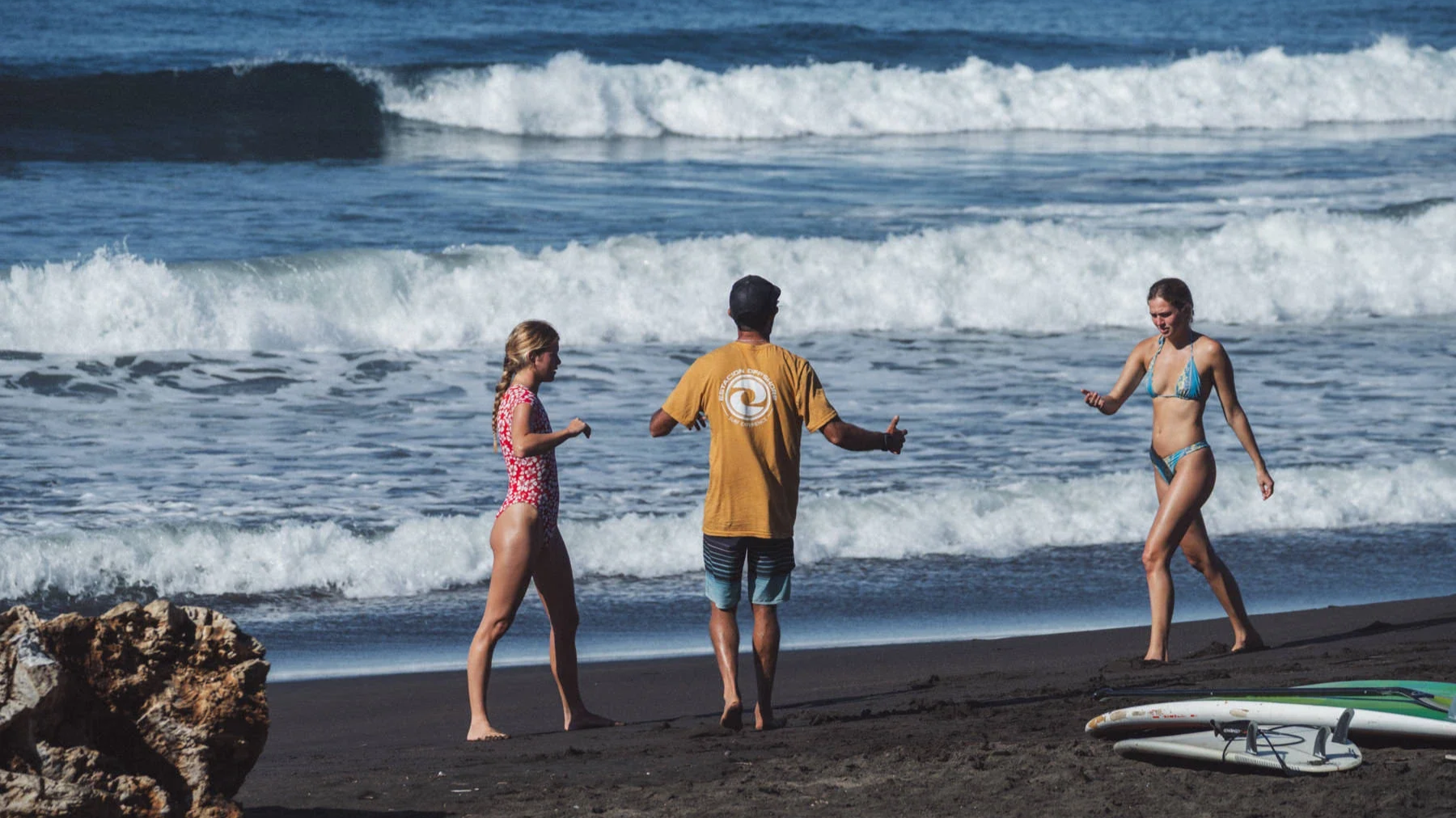An ISA-certified surf coach with two students during the lesson on the black sand beach of Playa Ostional Costa Rica, ocean waves and rocky outcrop in the background.