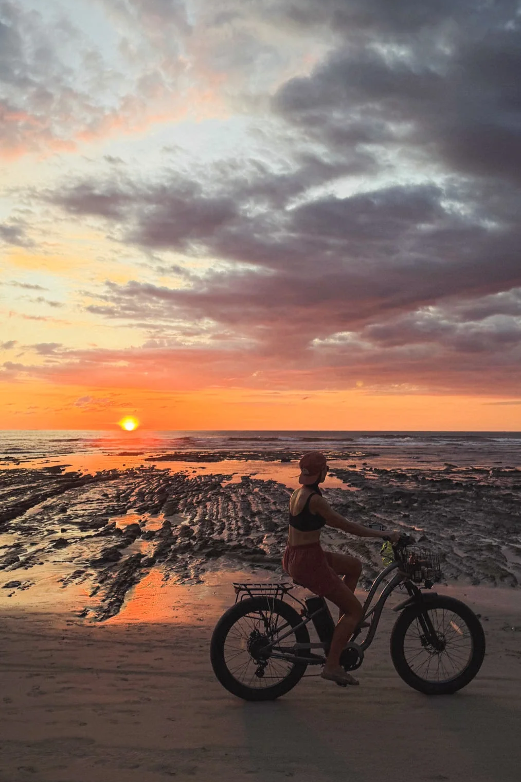 Woman cycling on the beach at Playa Guiones Nosara after a surf session, sunset sky with dramatic clouds overhead.
