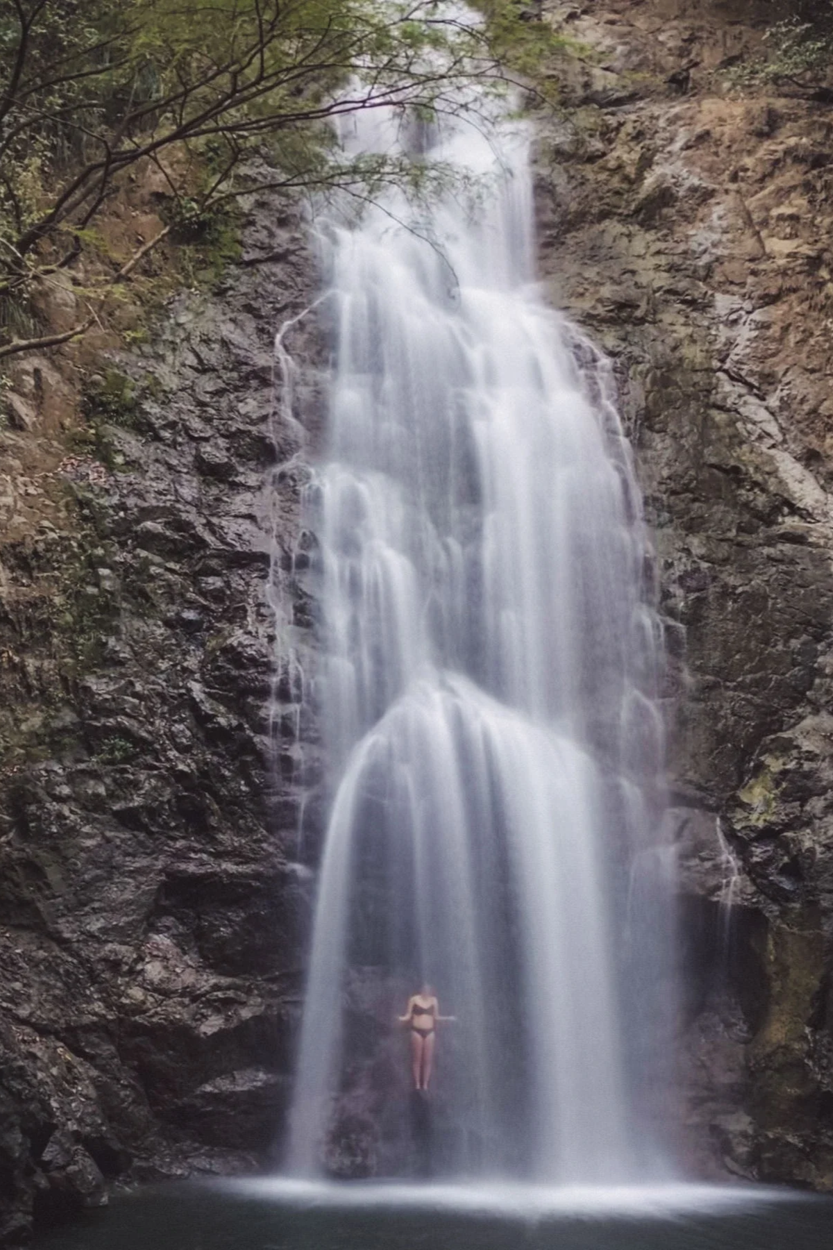A person in a black swimsuit standing under a tall waterfall in Costa Rica with water cascading down rocky cliffs into a pool below