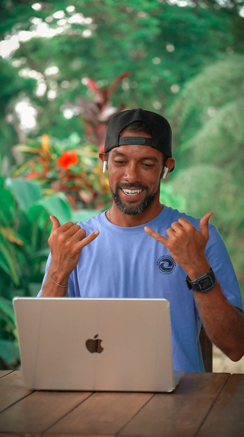 Surf instructor greeting during an online surf coaching video call on a laptop in a lush green outdoor setting in Costa Rica.