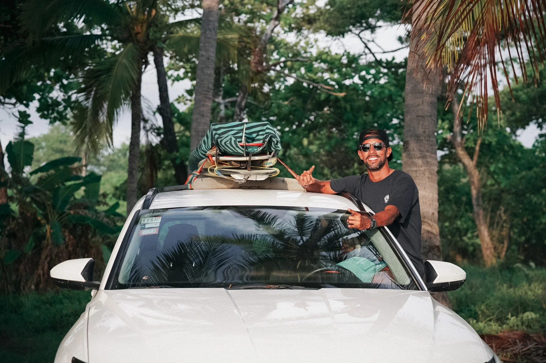Surf guide smiling and ready for a guided surf trip in Costa Rica, surfboards loaded on the roof of the car surrounded by tropical palm trees.